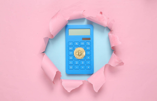 Калькулятор с биткойном через разорванную дыру на сине-розовом пастельном фоне. Концептуальное искусство. Пастельный цветовой тренд. Минимализм - Фото, изображение