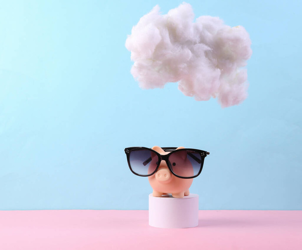 Sparschwein mit Sonnenbrille mit Wolke auf pastellrosa-blauem Hintergrund. Kreative Idee. Sommerpause. Minimales Konzept. Pastellfarbentrend - Foto, Bild