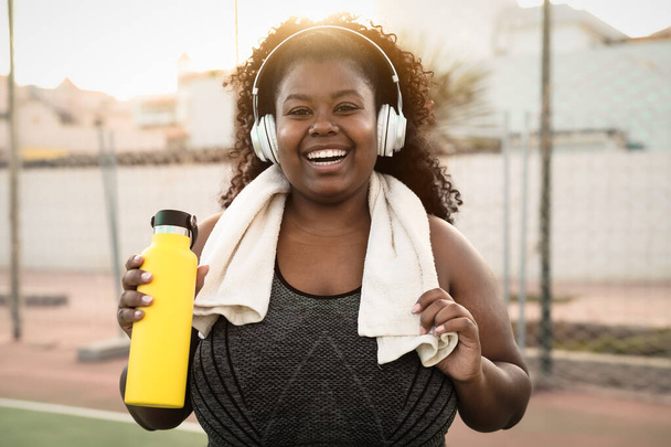 Feliz mujer africana con curvas haciendo jogging y rutina de ejercicios mientras escucha música con auriculares inalámbricos al aire libre - Foto, imagen