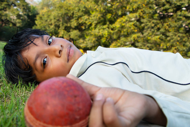 Мальчик лежит на земле и показывает крикетный мяч. - Фото, изображение