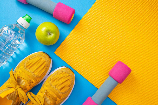 yellow sneakers and yellow mat, dumbbells, apple and water, blue background - 写真・画像