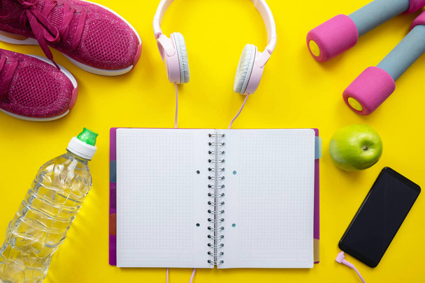 green apple and bottle of water, pink sneakers and dumbbells, notebook with pen and headphones with cell phone on yellow background - Photo, image