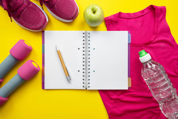 pink t-shirt, sneakers and dumbbells, notebook, apple and bottle of water on yellow background - Valokuva, kuva