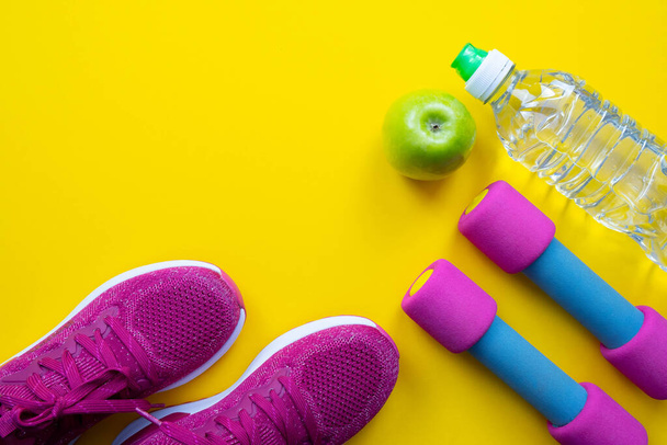 pink sneakers and dumbbells, green apple and bottle of water lie on the left side of the yellow background, close-up - Foto, Bild