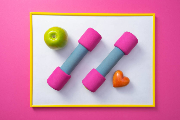 yellow frame with white background, pink dumbbells, green apple and heart, general pink background - Photo, image