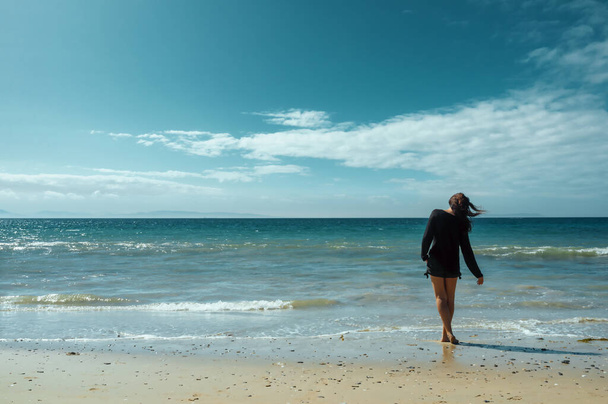 Mujer disfrutando de la cálida brisa en la playa vadeando en el oleaje en el borde de una playa tropical de arena con la brisa soplando su pelo - Foto, imagen