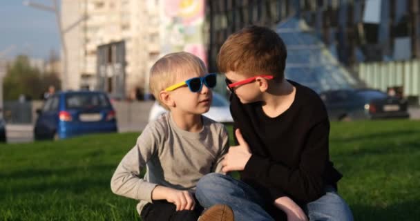 Zwei Jungs mit Sonnenbrille unterhalten sich auf dem Rasen. Kinder diskutieren munter etwas. Brüder ruhen sich auf dem Rasen aus - Filmmaterial, Video