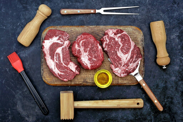 Μπριζόλες βοείου κρέατος για μπάρμπεκιου ή ψησίματος, Overhead View. Μπριζόλες βοδινού, Ξύλινο Σφυρί, Αλάτι και Πιπέρι στο ξύλο κοπής. Ακατέργαστο Striploin Marbled Beef Steaks σε μαύρο φόντο, πάνω όψη. - Φωτογραφία, εικόνα