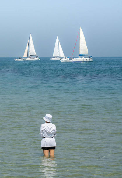 Een jonge vrouw gekleed in een witte top en hoed, staande in ondiep zeewater op een zonnige dag, kijken zeilboten gaan door. - Foto, afbeelding