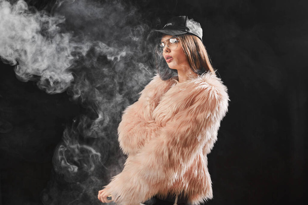 Sexy trendy rap woman in pink fur coat and cap smoke electronic cigarette. Pink Studio background shooting - 写真・画像