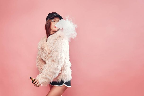 Sexy trendy rap woman in pink fur coat and cap smoke electronic cigarette. Pink Studio background shooting - Photo, Image