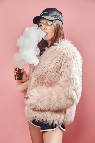 Sexy trendy rap woman in pink fur coat and cap smoke electronic cigarette. Pink Studio background shooting - Фото, изображение
