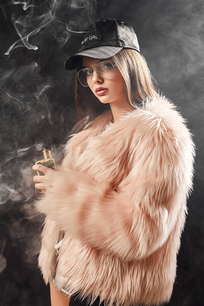 Sexy trendy rap woman in pink fur coat and cap smoke electronic cigarette. Dark Studio background shooting - Foto, Imagen
