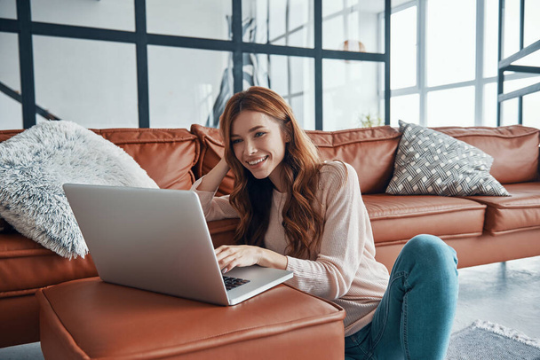 Attractive young woman using laptop and smiling while spending time at home - Φωτογραφία, εικόνα