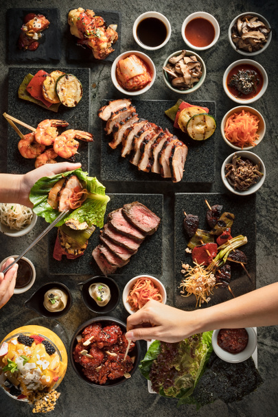 Flat lay of people preparing their Korean barbecue bites, they can select the meat such as beef and pork, there are also some shrimp or vegetable skewers, then they can add some pickled food, kimchi and wrap everything in salad leaf or nori wrap - Photo, Image