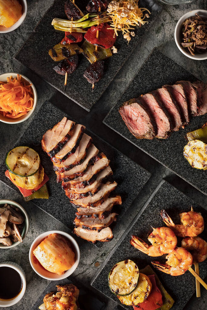 Nahaufnahme auf einem koreanischen Grill mit einer Auswahl an Rindfleisch, Schweinefleisch, Garnelenspießen, eingelegtem Gemüse und Sojasauce  - Foto, Bild