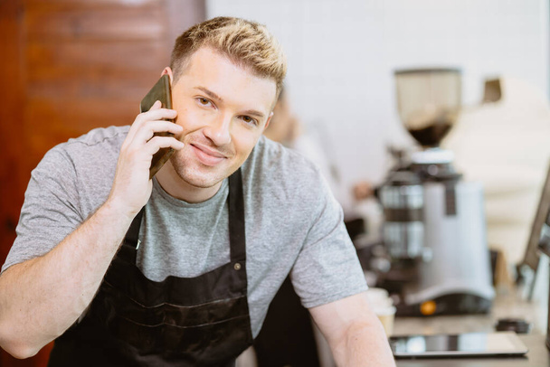 Barista mężczyzna dzwoni rozmawiać z klientem, aby otrzymać zamówienie napojów kawiarni przez telefon szczęśliwy uśmiech. - Zdjęcie, obraz