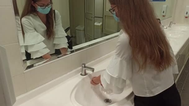 Η γυναίκα πλένει τα χέρια της στο νεροχύτη με αφρό. Έννοια της υγείας, τον καθαρισμό και την πρόληψη των μικροβίων και coronavirus από επαφή με τα χέρια. Μια κοπέλα με ιατρική μάσκα πλένει τα χέρια της σε μια δημόσια τουαλέτα. - Πλάνα, βίντεο