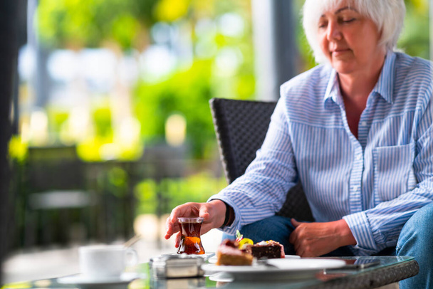 Woman enjoying a mug of hot tea and cakes on a patio of an outdoor restaurant in a cropped view as she reaches for the beverage with copyspace - Valokuva, kuva