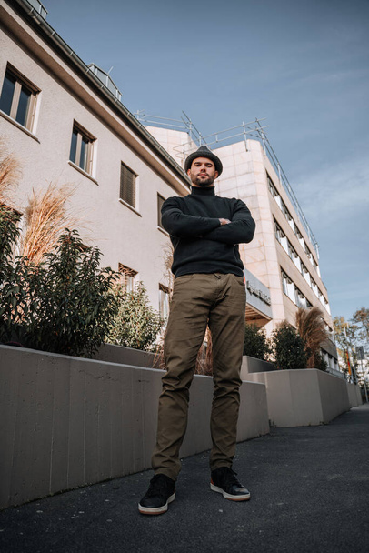 A young man in dark pants, black sweater and a hat, with arms crossed in front of a municipal building, upward view - Foto, Imagen