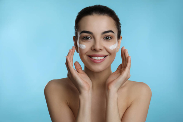 Young woman applying facial cream on light blue background - Foto, Imagen
