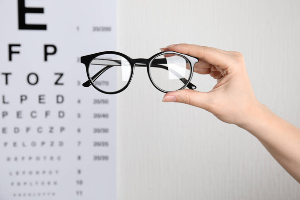 Woman holding glasses against eye chart on light background, closeup. Ophthalmologist prescription - Photo, Image