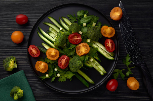 A black round plate of fresh vegetables stands on a black wooden table with a black knife, tomatoes and broccoli nearby. Top view.  - Foto, Imagen