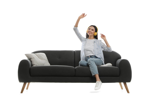 Mujer joven escuchando música en un cómodo sofá gris sobre fondo blanco - Foto, Imagen