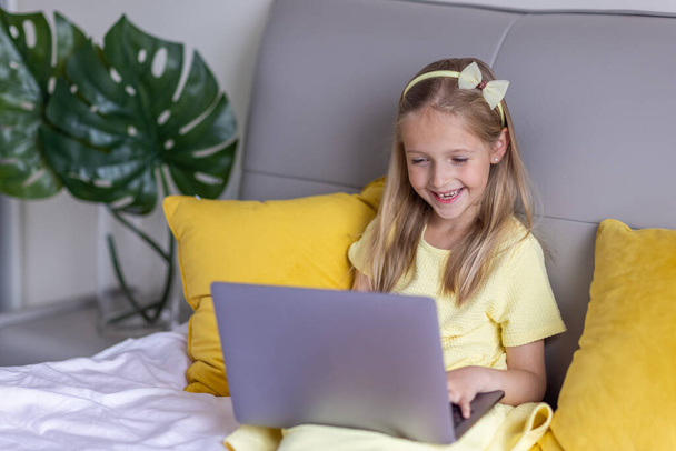 Cute little caucasian girl with blonde hair in fashionable dress illuminating yellow color sitting at home during coronavirus pandemic quarantine and using laptop. Stay at home during covid-19 - 写真・画像