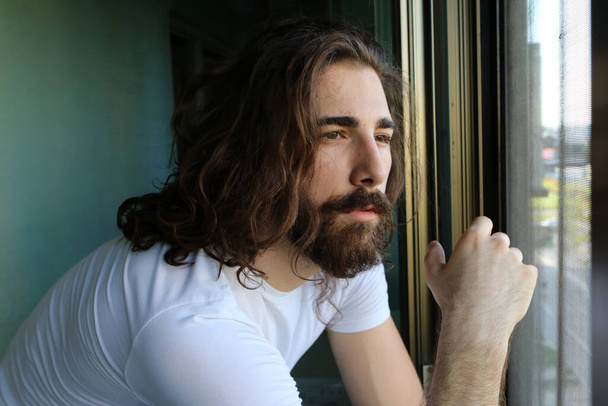 Стильный молодой кавказский парень с длинными волосами и бородой позирует на балконе. Стиль, тенденции, концепция моды. - Фото, изображение