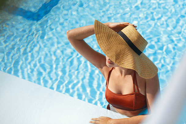 Beautiful woman in hat relaxing in pool water, enjoying summer vacation at tropical resort. Slim young female sunbathing at swimming pool edge, view above. Holidays and travel. Space for text - Foto, Imagen