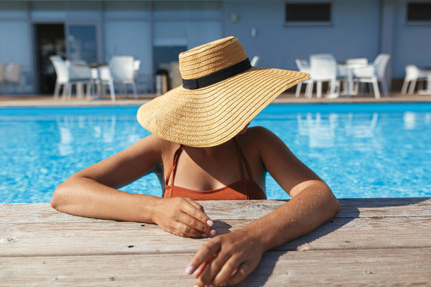 トロピカルリゾートで夏休みをお楽しみください。美しい女性の帽子プール木製の桟橋で水でリラックス。スイミングプールの端でスリム若い女性の日光浴、上記のビュー。休日と旅行 - 写真・画像
