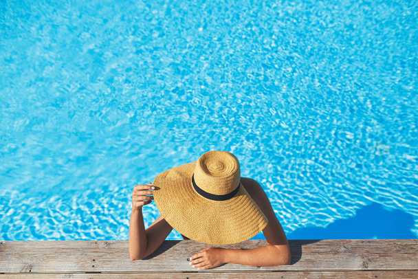 夏休みを楽しんで。木製の桟橋でプールの水でリラックスした帽子のスタイリッシュな女性、トップビュー。水泳プールの端でスリムな若い女性の日光浴。旅行や休日。テキストのスペース - 写真・画像