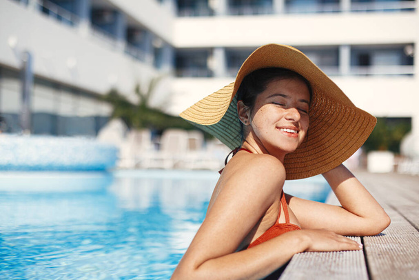 Enjoying summer vacation. Beautiful happy woman in hat relaxing in pool water. Stylish slim young female sunbathing and resting at swimming pool edge at tropical resort. Holidays and travel - Photo, image