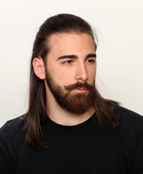 Joven modelo masculino de moda con el pelo largo y la barba posando en el estudio sobre un fondo aislado. Moda, negocio, concepto de modelaje. - Foto, imagen