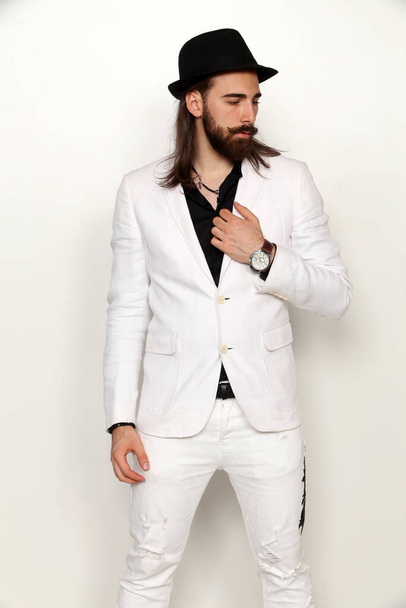 Молодая, модная мужская модель с длинными волосами и бородой позирует в студии на изолированном фоне. Мода, бизнес, концепция моделирования. - Фото, изображение