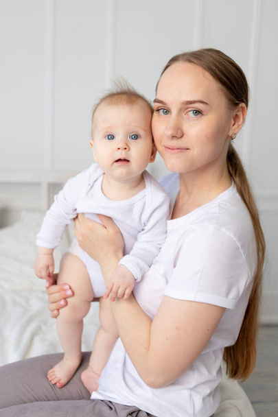 πορτρέτο μιας μητέρας με ένα μωρό στην αγκαλιά της αγκαλιάζοντάς το απαλά σε ένα λευκό κρεβάτι στο σπίτι, η αγάπη της μητέρας - Φωτογραφία, εικόνα