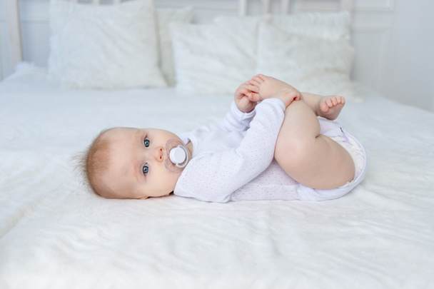 um bebê com uma chupeta por seis meses em uma cama branca encontra-se em suas costas e mantém suas pernas - Foto, Imagem