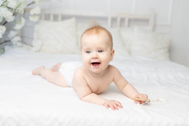 happy smiling baby in diapers on a white cotton bed at home six months old - Photo, Image