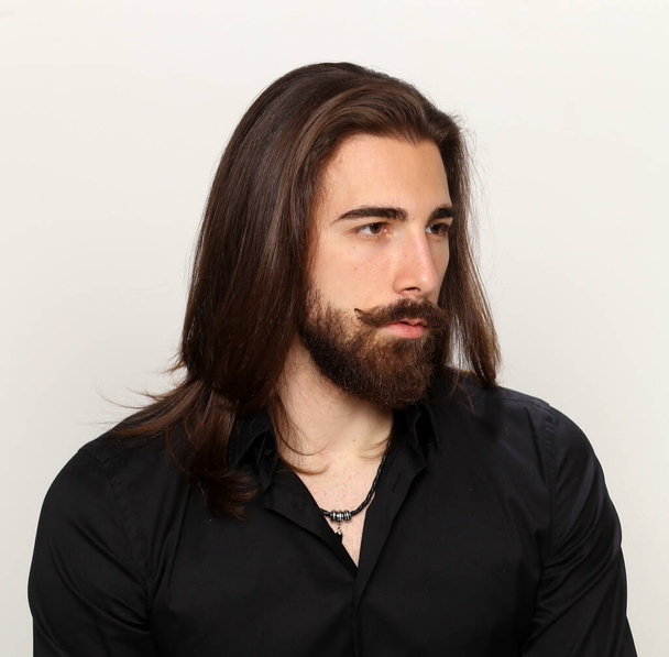 Jeune mannequin masculin à la mode aux cheveux longs et à la barbe posant en studio sur fond isolé. Mode, affaires, concept de modélisation. - Photo, image