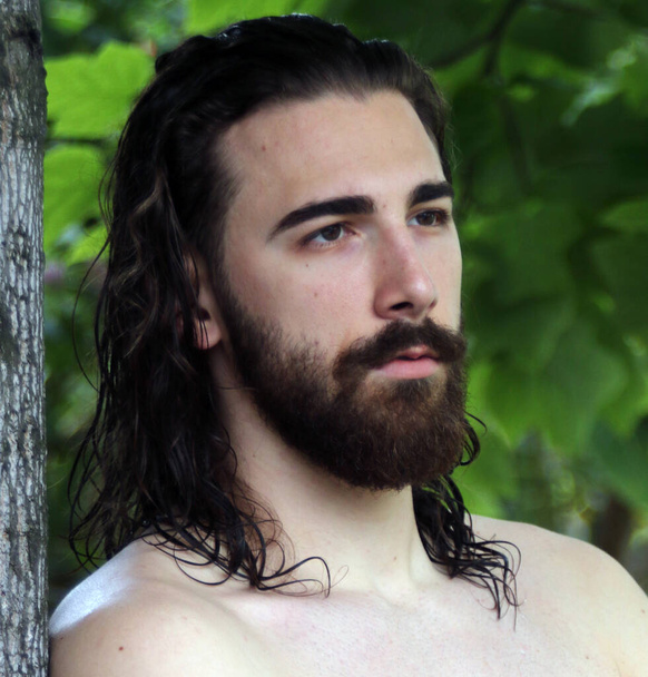Mladý atraktivní mužský model s dlouhými vlasy a vousy pózující v přírodě. Styl, trendy, módní koncept. - Fotografie, Obrázek