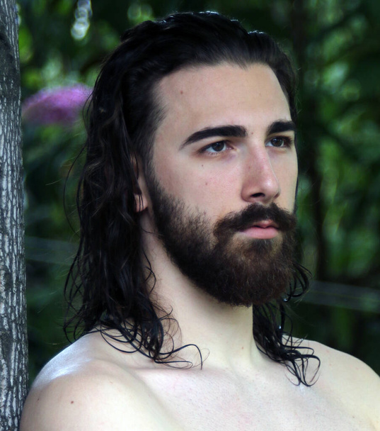 Молодой, привлекательный мужчина-модель с длинными волосами и бородой позирует в природе. Стиль, тенденции, концепция моды. - Фото, изображение
