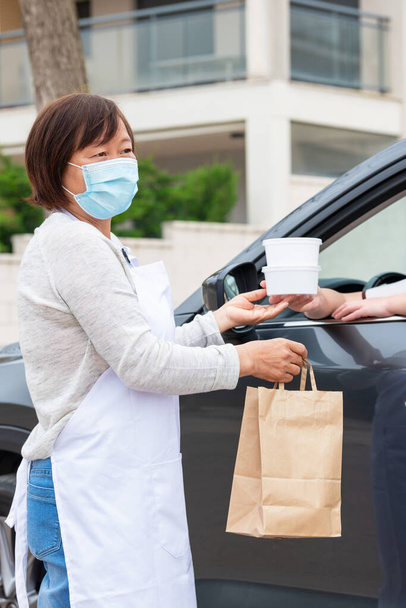 Aasialainen nainen toimittaa noutoruokaa asiakkaalle autossa: Valikoiva tarkennus. Liiketoiminnan ja turvallisuuden käsite. - Valokuva, kuva