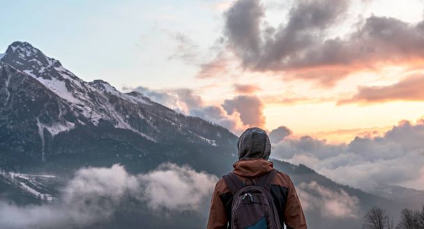 Young man traveler in hood with backpack from behind looking mountain view of the snow capped peaks of Caucasus Mountains in clouds at sunset - Photo, Image
