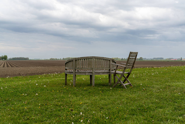 Prázdný dřevěný zahradní nábytek na zeleném poli pod zataženou oblohou s čerstvě oraným polem za ním - Fotografie, Obrázek