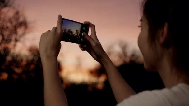 Νεαρή ευτυχισμένη ασιατική κορίτσι κατέχουν κινητό τηλέφωνο χαλαρώσει χαρούμενα τραβήξτε φωτογραφία ειρηνική εικόνα του ηλιοβασιλέματος στο δάσος δέντρο στο πάρκο. Όμορφη θέα στη φύση ηλιοβασίλεμα, διακοπές ταξιδιού με την έννοια smartphone. - Πλάνα, βίντεο