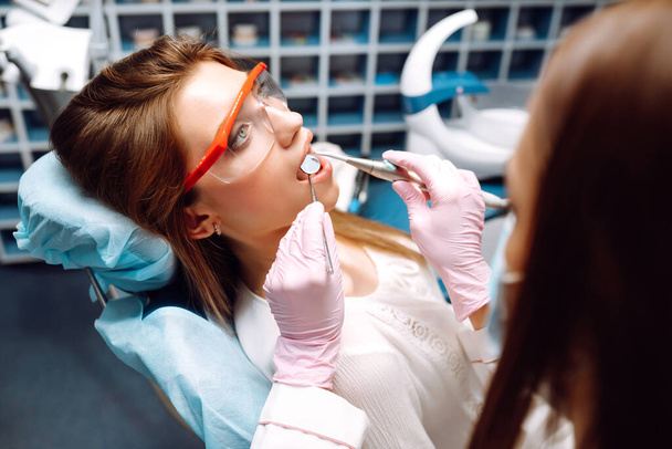 Young woman opening her mouth wide during treating her teeth by the dentist. Hands of a doctor holding dental instruments near patient's mouth. Healthy teeth and medicine concept. - 写真・画像