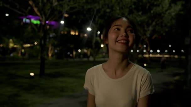 Fiatal boldog ázsiai lány pihenni séta a gyalogúton és vidám mosoly nézi kamera körül fa a parkban a város éjszakáján. Gyönyörű természet a városra néző, Utazás nyaralás nyaralás tevékenység koncepció. - Felvétel, videó