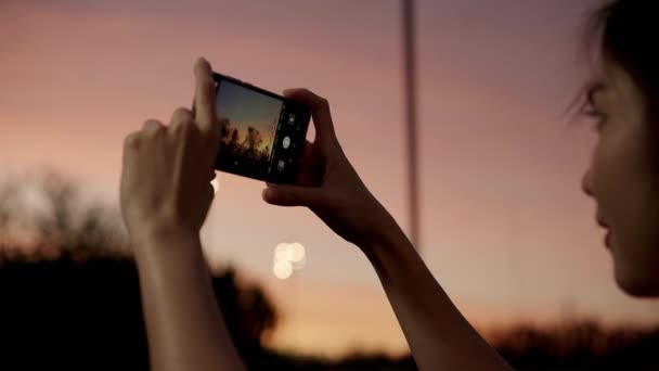 Young happy asian girl hold mobile phone relax cheerful take photo peaceful picture of sunset at forest tree in park. Beautiful nature sunset view, Travel holiday vacation with smartphone concept. - Video