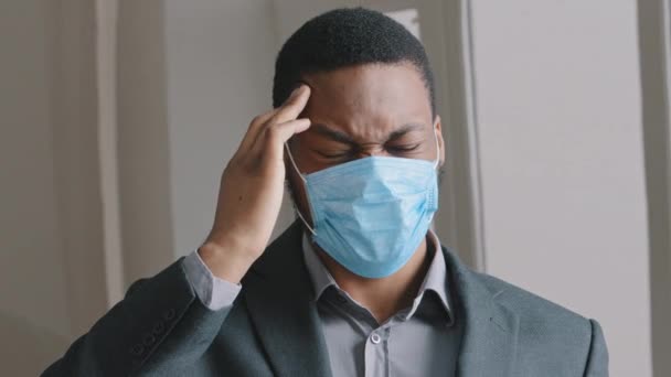 Face of unhappy young african american businessman. Concerned young man wears face mask rubs temple, closed eyes suffers headache painful feelings migraine hurt. Feeling tired, suffering from covid - Footage, Video
